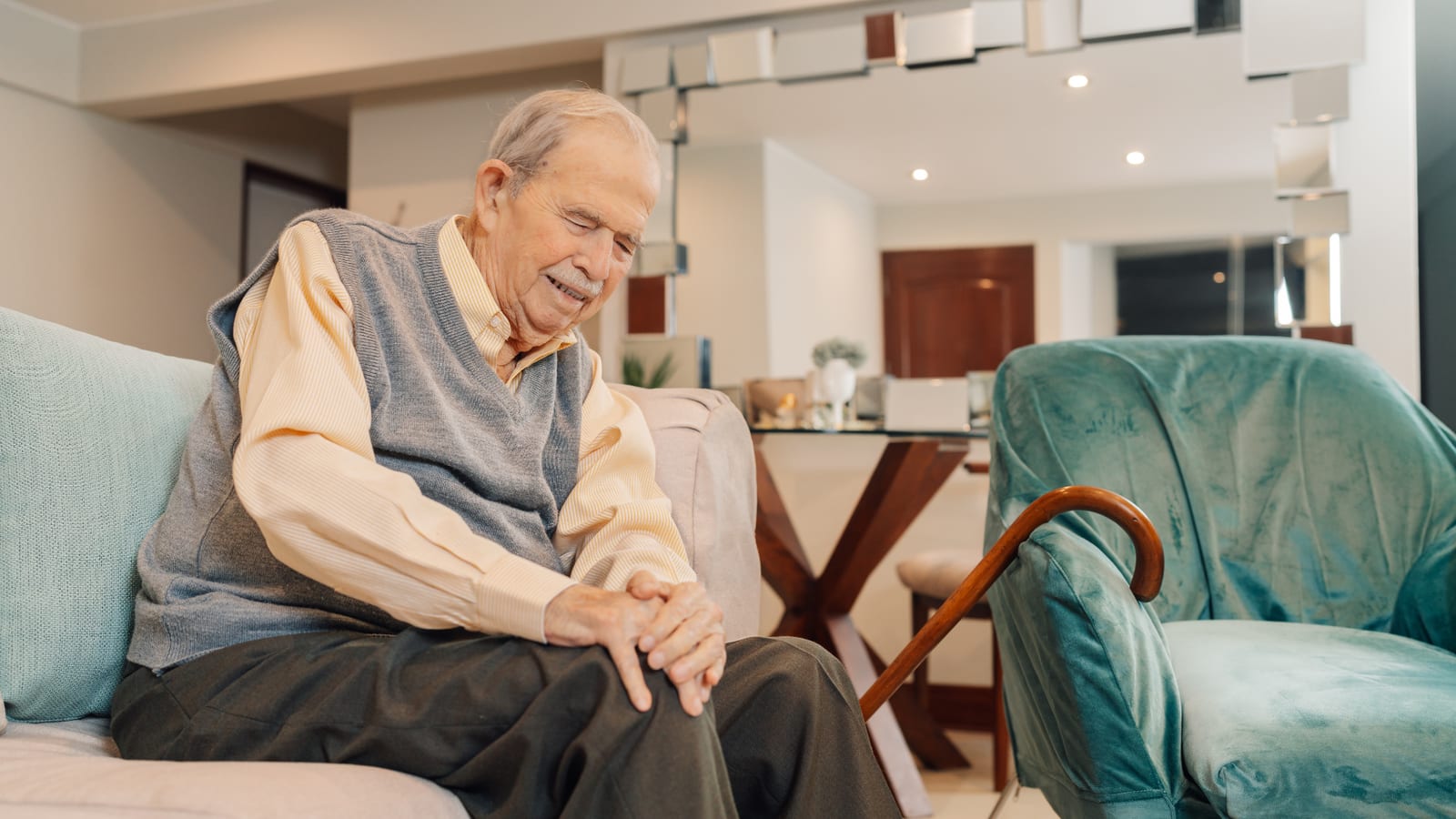 Older man sitting alone on a couch at home with his hands folded and a cane resting beside him