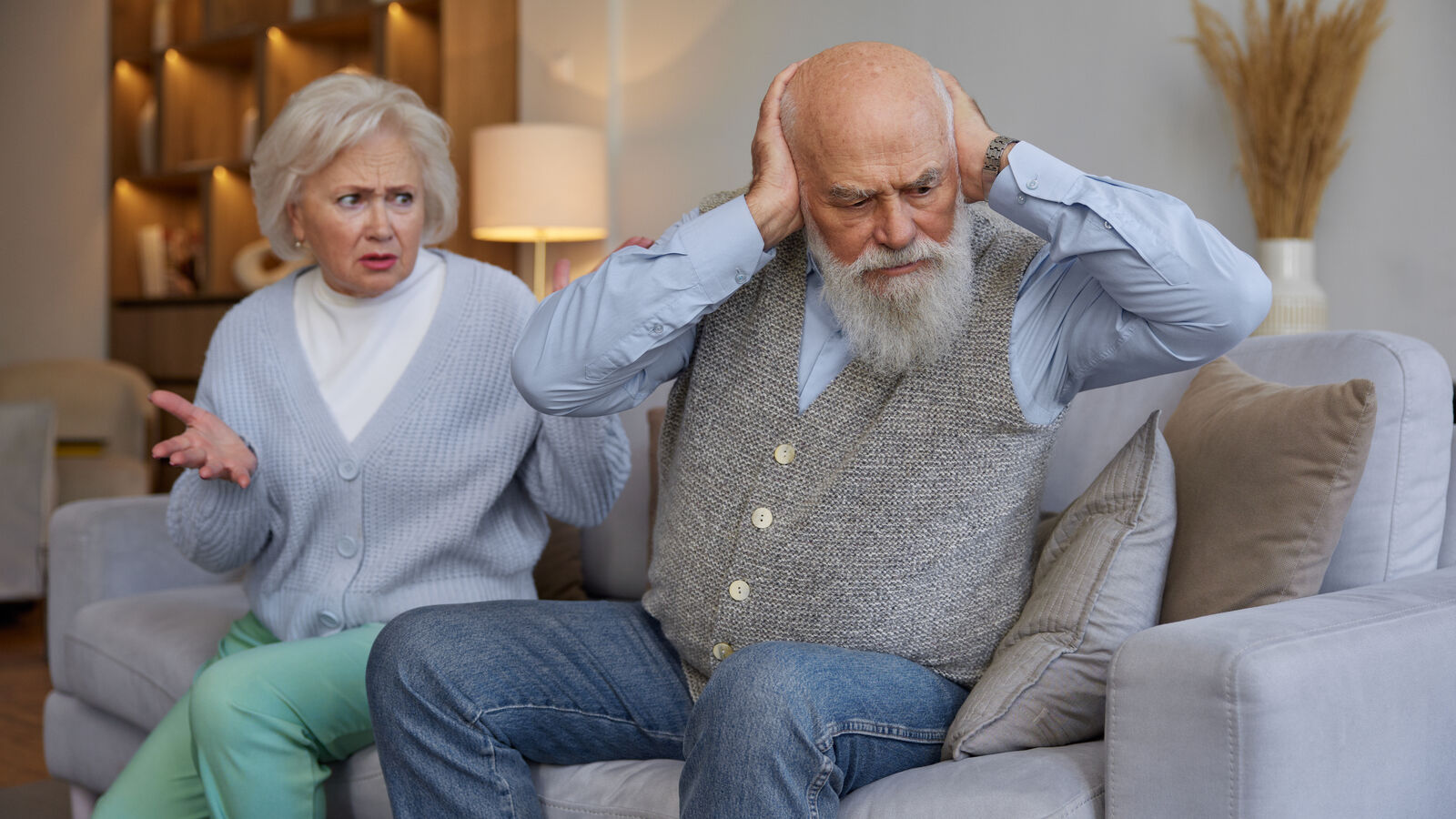 Older man on a living room couch covering his ears while his wife sits behind him looking frustrated and worried