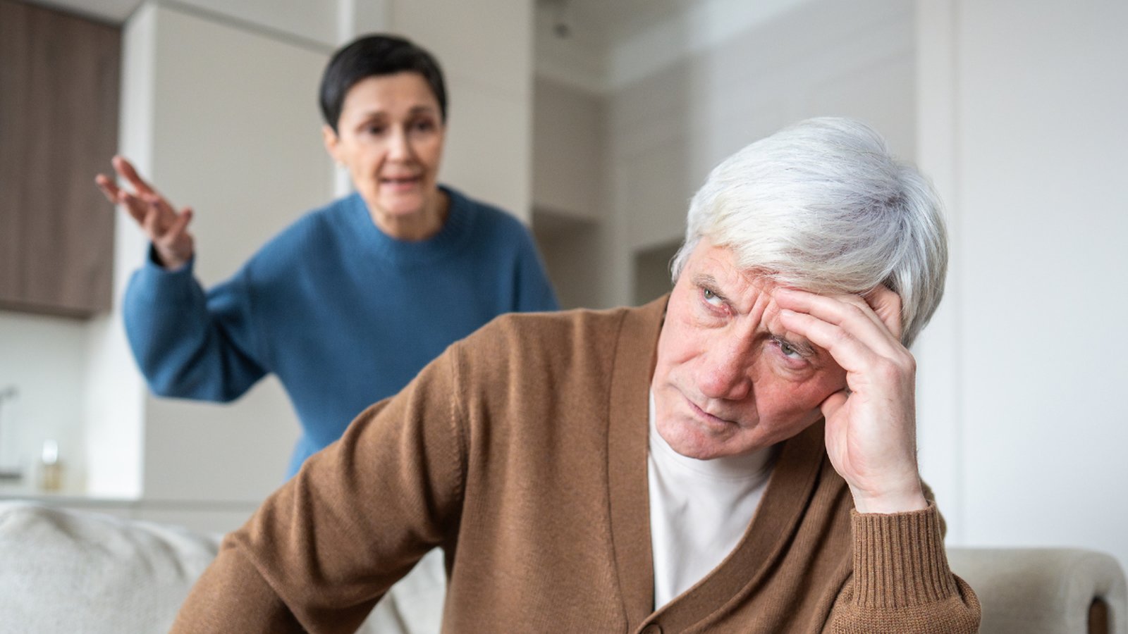 Older man sitting on a couch with his head in his hand while a family member stands behind him mid-conversation